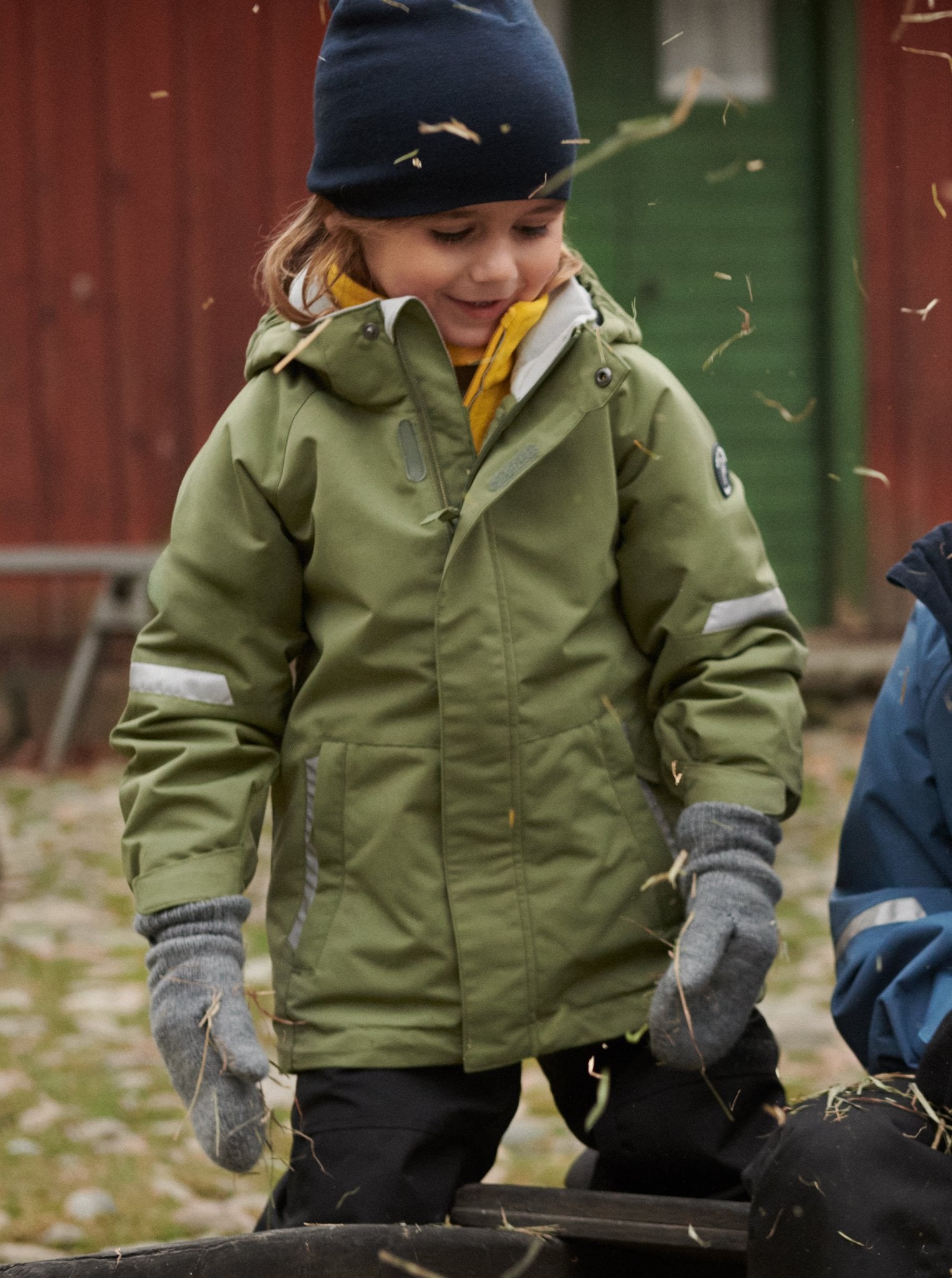Girl playing outside wearing 100% waterproof kids coat in green and 100% waterproof trousers in black. Accessories with warm merino wool beanie hat in navy and grey wool gloves