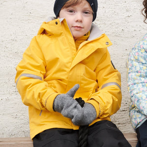 Boy sitting outside wearing 100% waterproof kids coat in bright yellow and 100% waterproof trousers in black. Accessories with warm merino wool beanie hat in navy