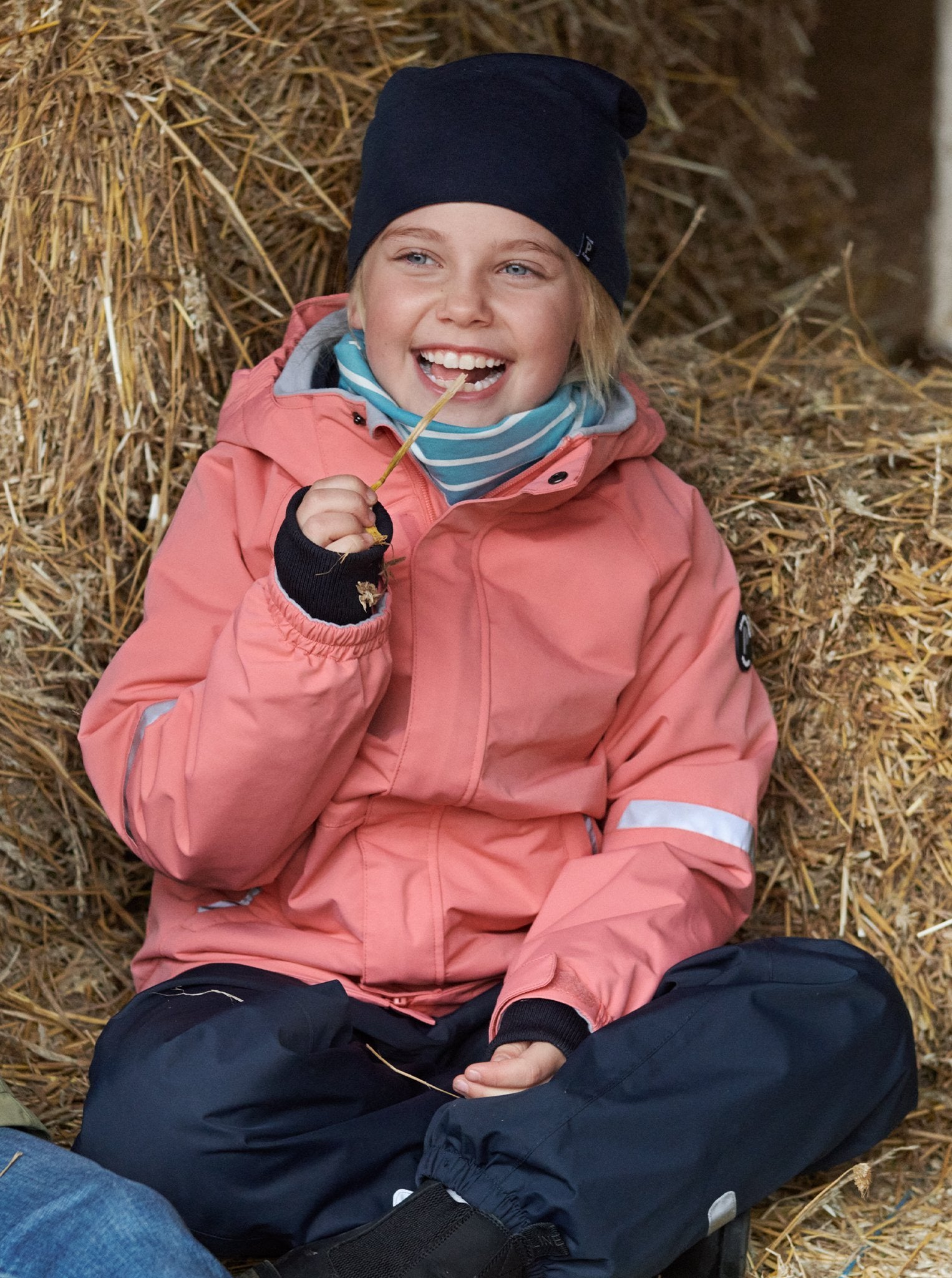 Girl sitting in hay barn wearing 100% waterproof kids coat in pink and 100% waterproof trousers in black. Accessories with warm merino wool beanie hat in navy and blue and white stripe organic cotton tube scarf 