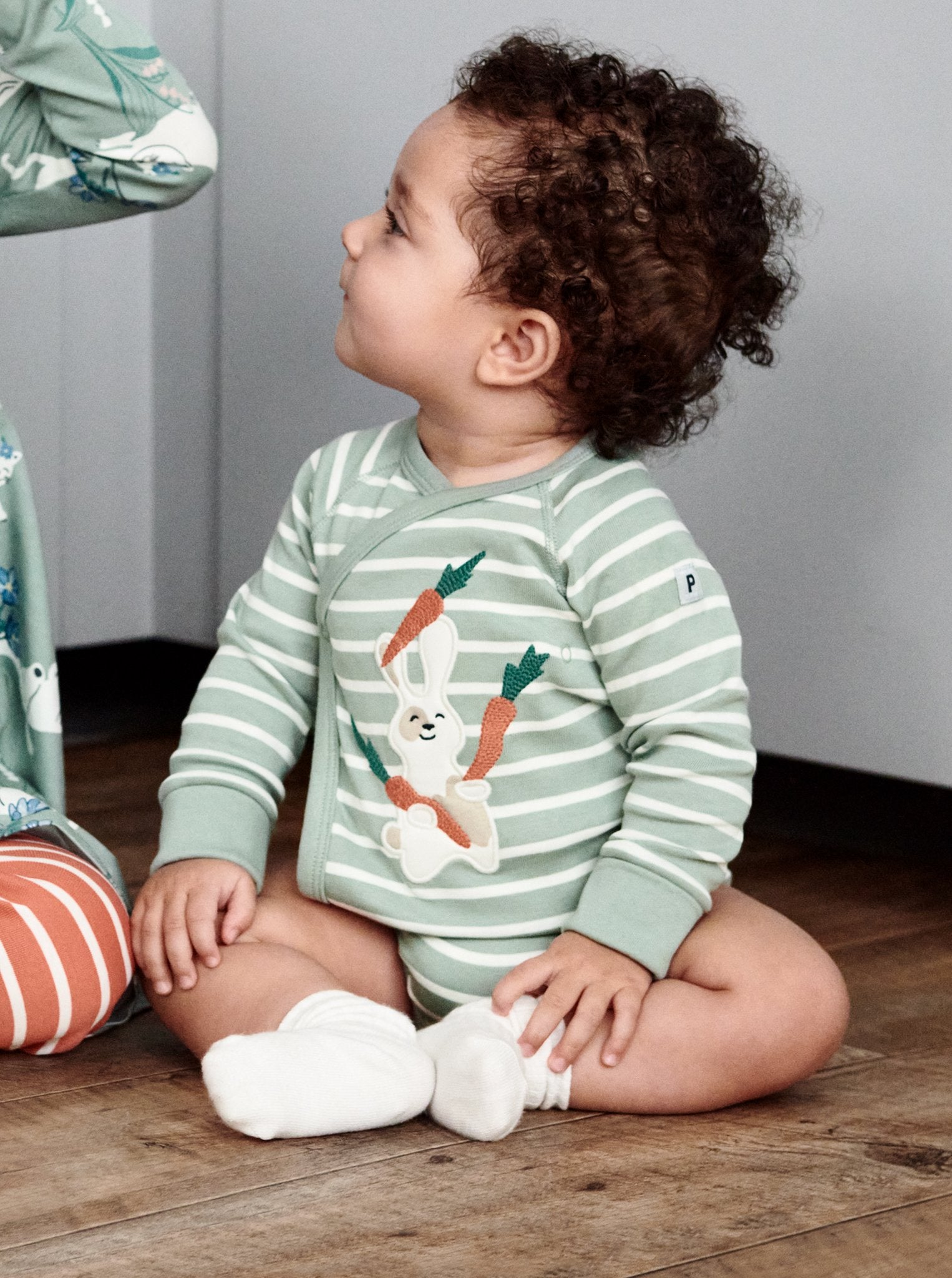 A toddler wearing a green and white striped cotton long sleeve babygrow with fun bunny eating carrots applique, paired with white organic cotton socks