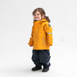 A little girl wearing a yellow, kids waterproof jacket, paired with black, kids waterproof trousers, made of shell fabric.