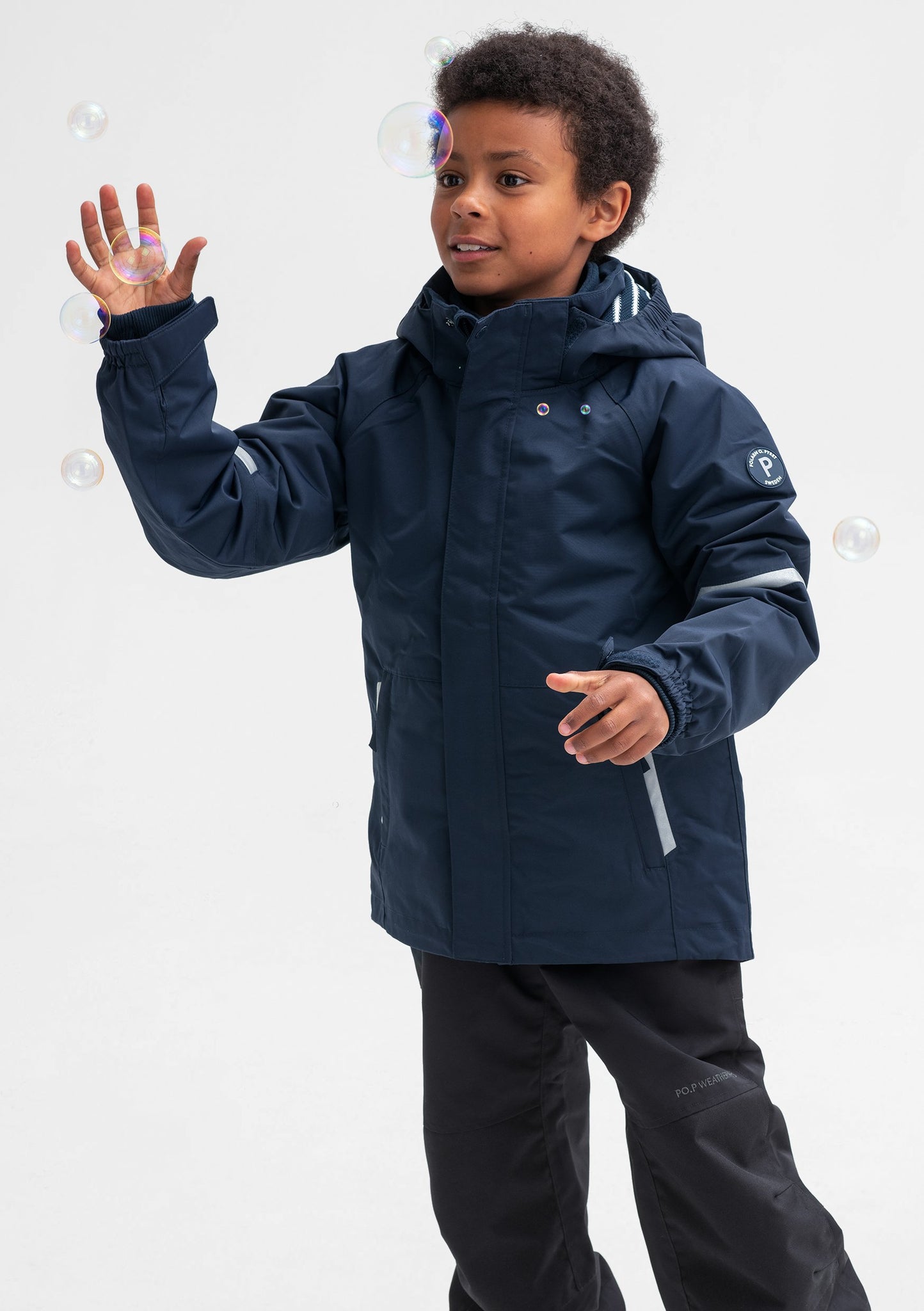 A little boy sporting a waterproof kids jacket in navy, with detachable hood and reflectors, made of soft shell fabric.