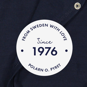 White round label with the text 'From Sweden With Love, Since 1976" shown in front of a navy blue babygrow background.